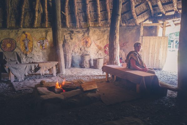 Woman dressed in Iron Age costume sitting inside roundhouse as campfire burns