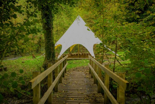 White tipi viewed from across a wooden bridge with trees and grass surround it