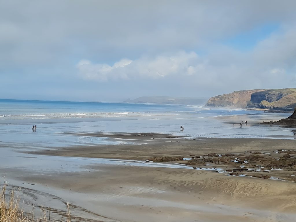 View across a sandy beach with a handful of people walking across the wet sand as the tide retreats on a bright day with white clouds dominating the blue sky. Location featured is Little Haven, Pembrokeshire