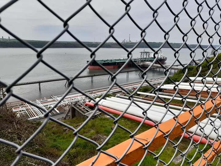 View of a ship docked at an industrial facility, photograph taken through a chain link fence.