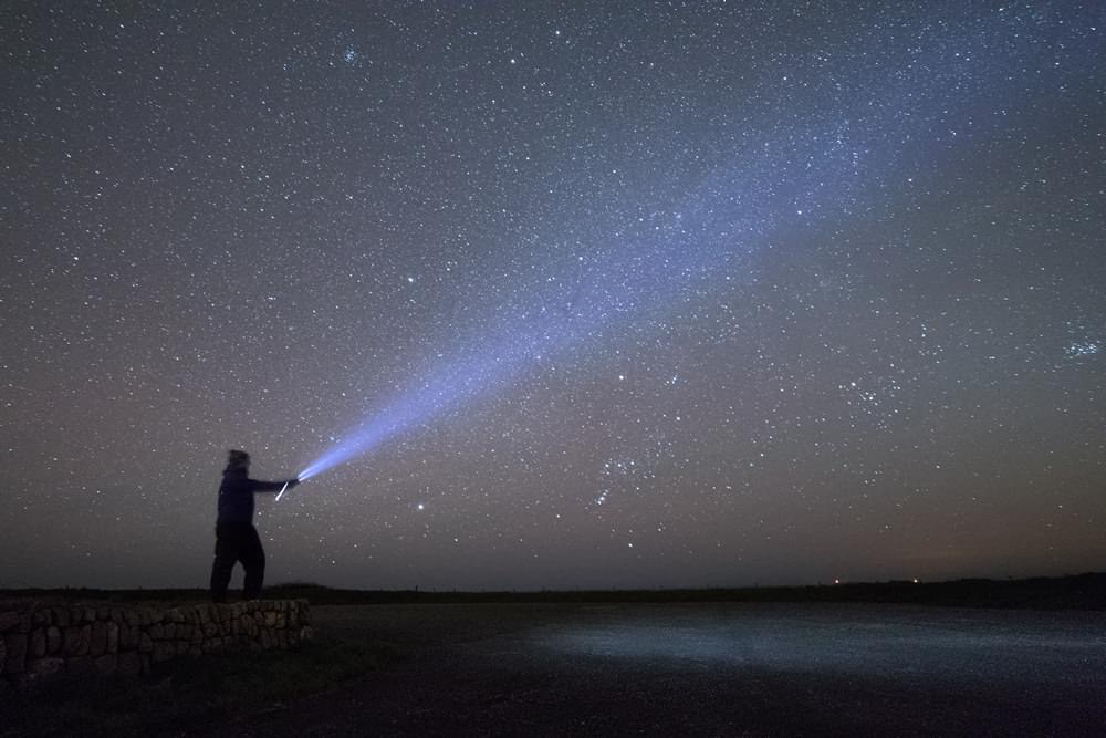 A silhouette of a person beaming a flashlight towards a starry night sky